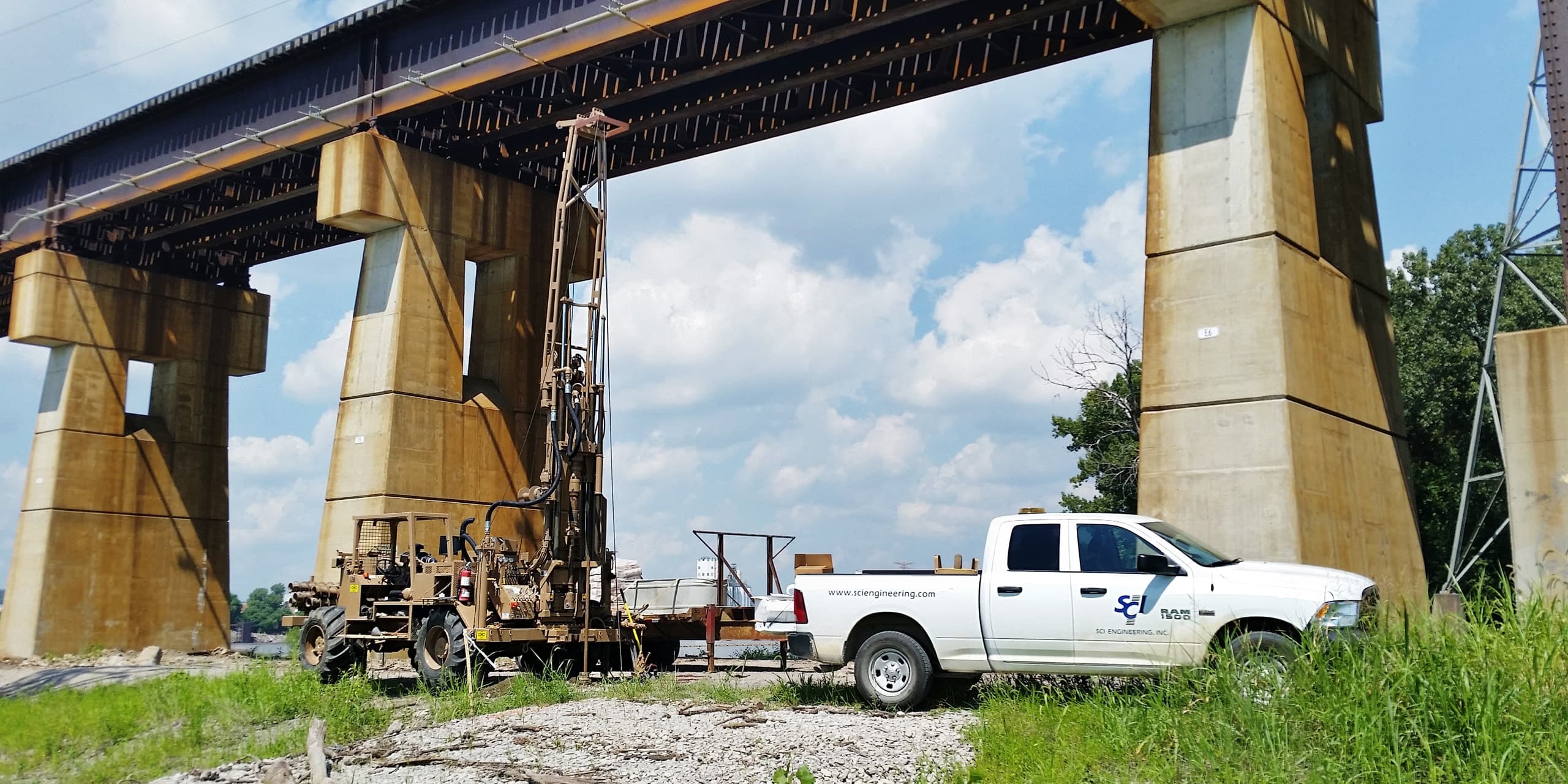An SCI Engineering drill rig and company pickup at a project site beneath a river bridge
